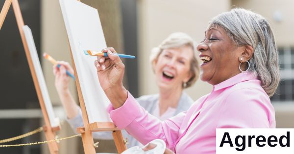 One woman paints a painting while another looks on, both smiling.