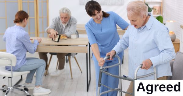 A health care worker helps a woman using a walker while two people talk at a table behind them.